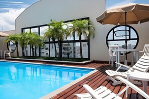 A pool area with a wooden deck, lounge chairs, and a table with an umbrella. - Flat Moderno Proximo ao Aeroporto (São Paulo)