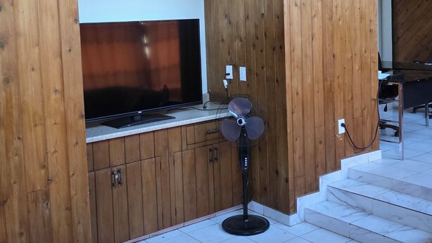 Quarto com paredes de painel de madeira, TV de tela plana, ventilador de chão e escada que leva a um andar superior.