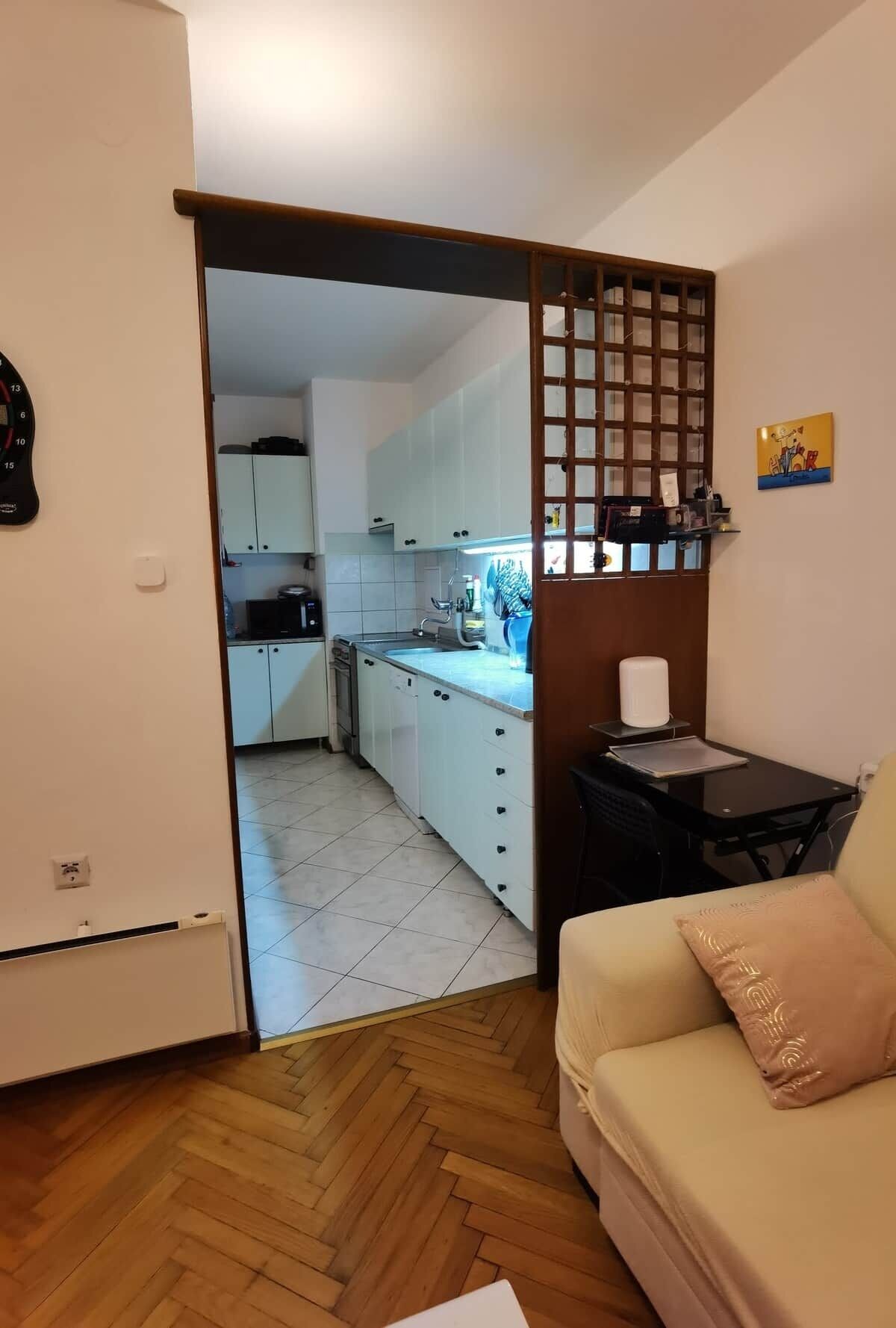 Un espacio habitable compacto con kitchenette, piso de madera en patrón de espiga, sofá color beige y una pequeña mesa.