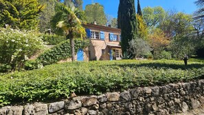 A two-story house with a stone wall in the foreground, surrounded by lush greenery and trees. - Charming Provençal villa with a pool and sauna (MONTFERRAT)