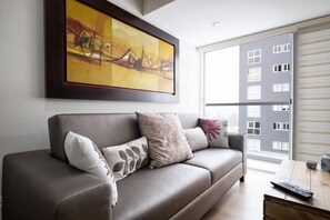 A modern living room with a grey sofa, a large abstract painting, and a view of a building through a window. - Your Furnished Apartment on Pardo Avenue, Miraflores, Lima – Excellent Location (Miraflores)