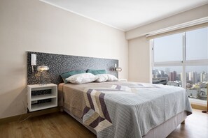 Ein Schlafzimmer mit einem großen Bett, zwei Nachttischlampen, einem Nachttisch, einer eingebauten Ablage und Blick auf die Stadt durch ein großes Fenster.