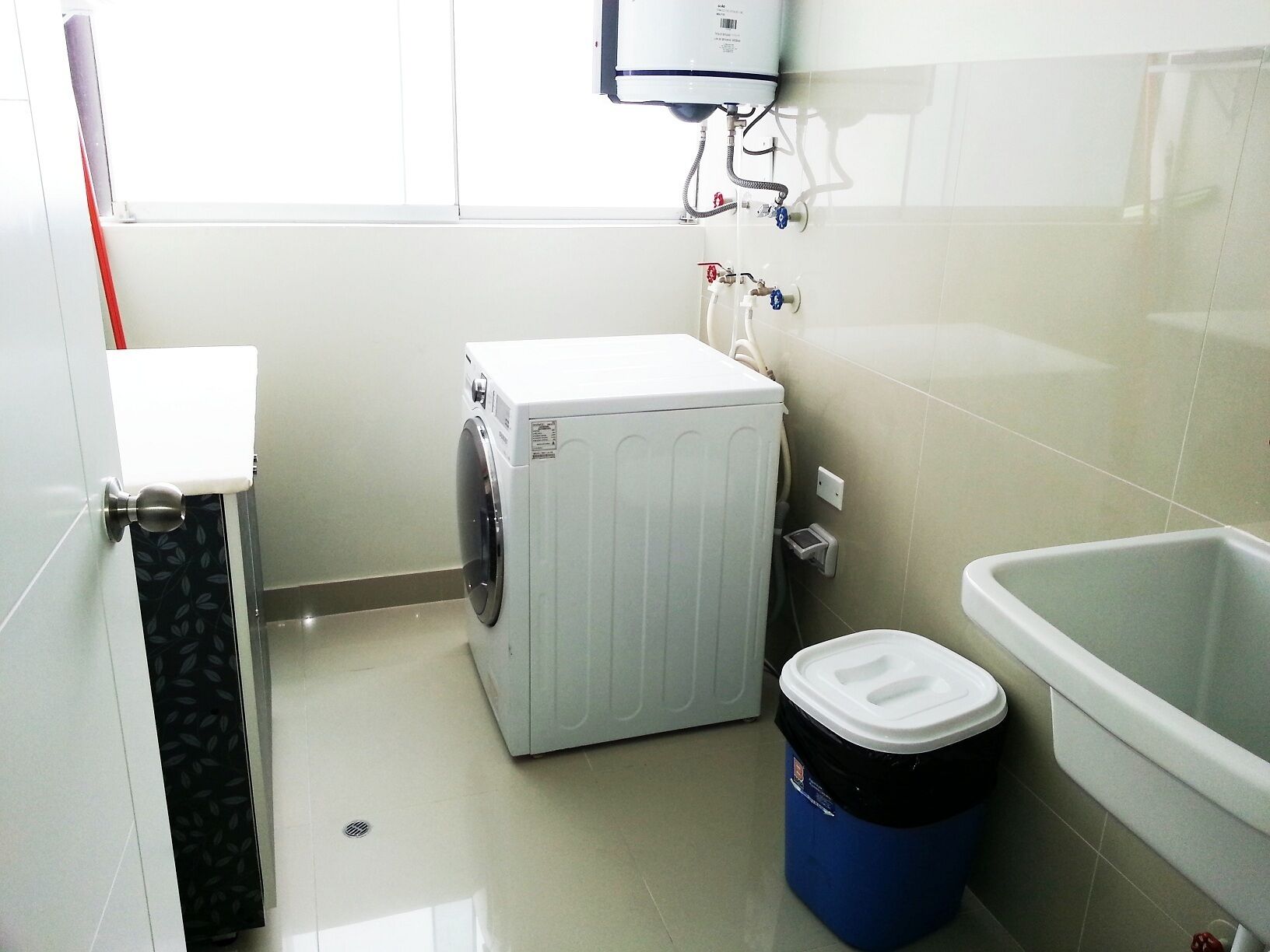 A compact laundry room with a washing machine, a trash bin, and a bathtub.