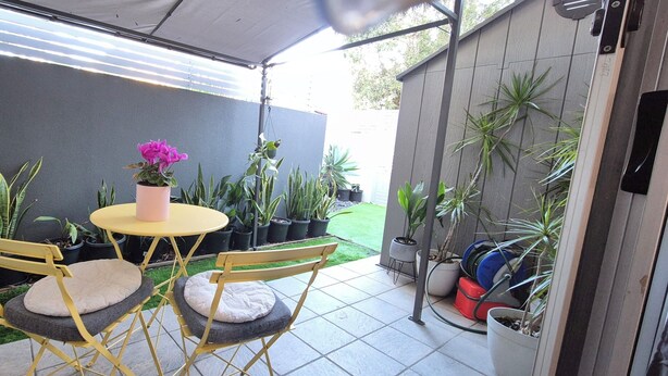Een terras met een gele tafel en stoelen, omringd door potplanten en een klein gazon.