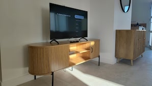 A modern living room with a flat-screen TV mounted on a wooden console, a wooden cabinet with drawers, and a mirror on the wall. - Quiet house just 5 minutes from the beaches with direct access to the bike path  (Clohars-Carnoët)