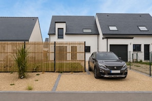 A modern two-story house with a car parked in the driveway, a wooden fence, and a gravel path.