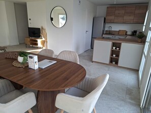Un espai de vida modern amb una taula de menjador de fusta, cadires blanques amb coixins, una zona de cuina amb armaris de fusta i un mirall gran a la paret.
