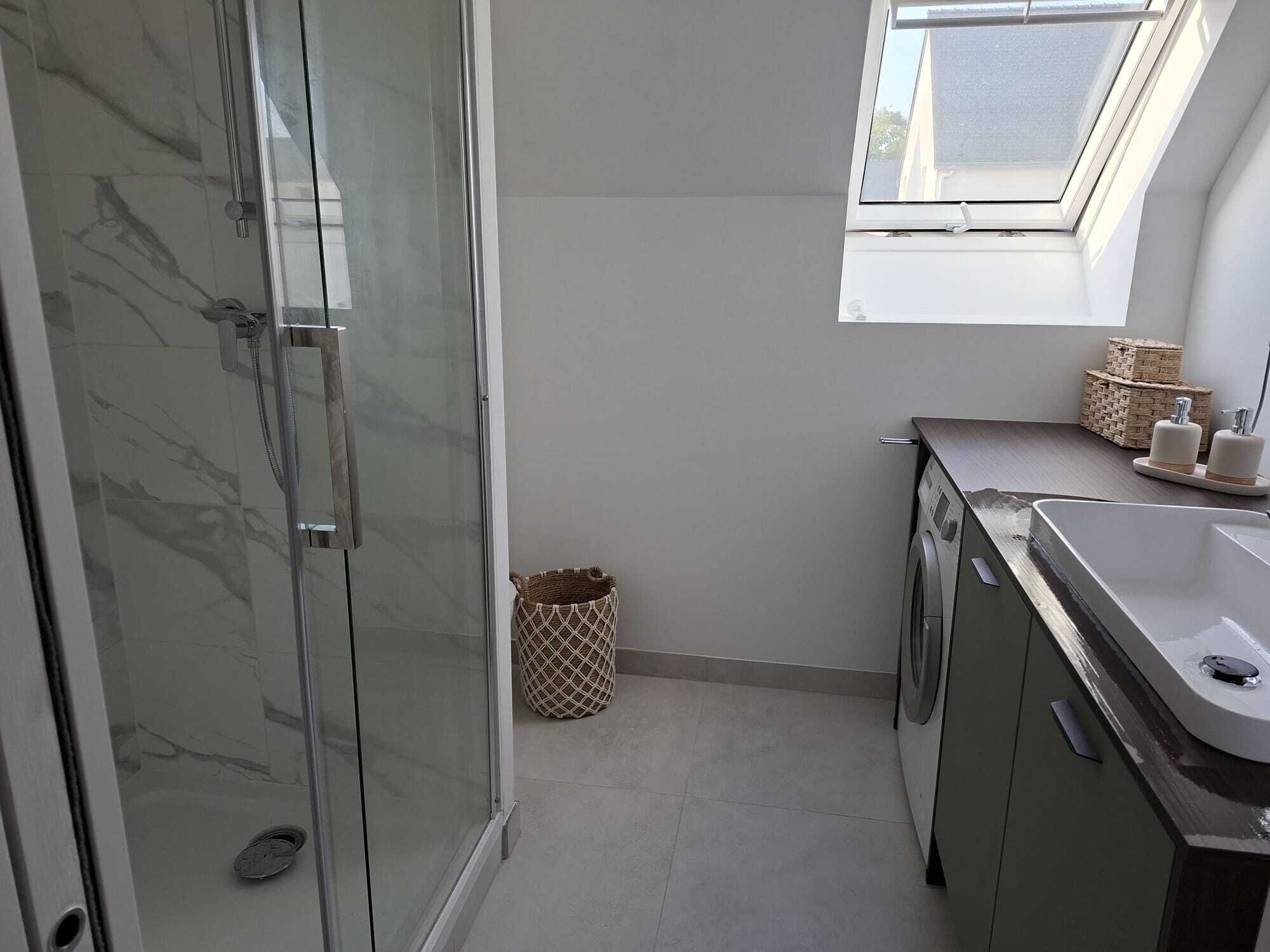 Une petite salle de bain dotée d’une douche à parois de verre, d’un lave-linge, d’un lavabo et d’une petite fenêtre.