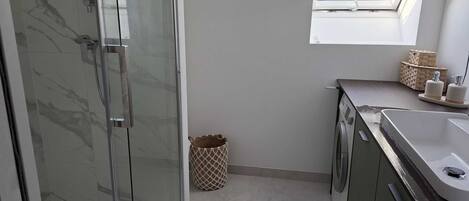 Une petite salle de bain dotée d’une douche à parois de verre, d’un lave-linge, d’un lavabo et d’une petite fenêtre.