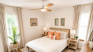 A bedroom with a bed, nightstands, a ceiling fan, a window with curtains, a potted plant, and framed artwork on the walls. - The Willow-A Seabrook Coastal Retreat Near Beaufort & Hunting Island (Seabrook)