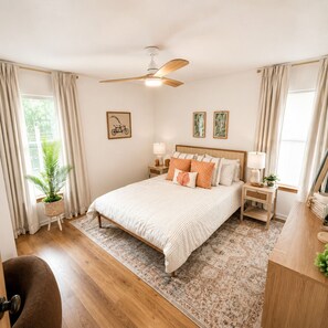 Une chambre à coucher comprenant un lit, des tables de chevet, un ventilateur de plafond, une fenêtre avec des rideaux, une plante en pot et des œuvres d’art encadrées accrochées aux murs.