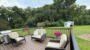 A deck with wicker furniture, a grill, and a small shed in a wooded area. - The Willow-A Seabrook Coastal Retreat Near Beaufort & Hunting Island (Seabrook)