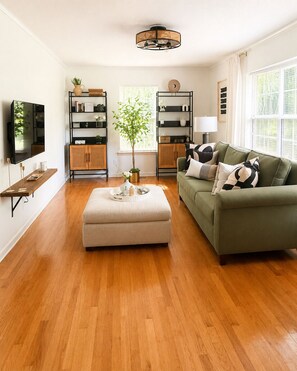 Un salon avec un canapé vert, un pouf blanc et une table basse en bois. On y trouve deux étagères noires ornées d’objets décoratifs, un téléviseur fixé au mur et une fenêtre avec des rideaux blancs.