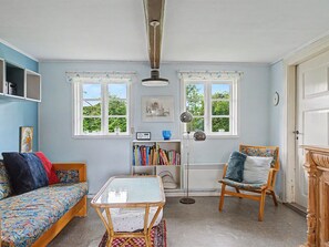 Una habitación con un sofá, una silla, una mesa, una estantería y dos ventanas.