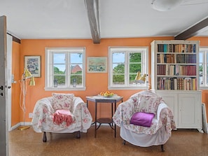 Una habitación con dos sillones estampados, una mesita baja con fruta, una estantería y ventanas amplias con vistas al verdor.