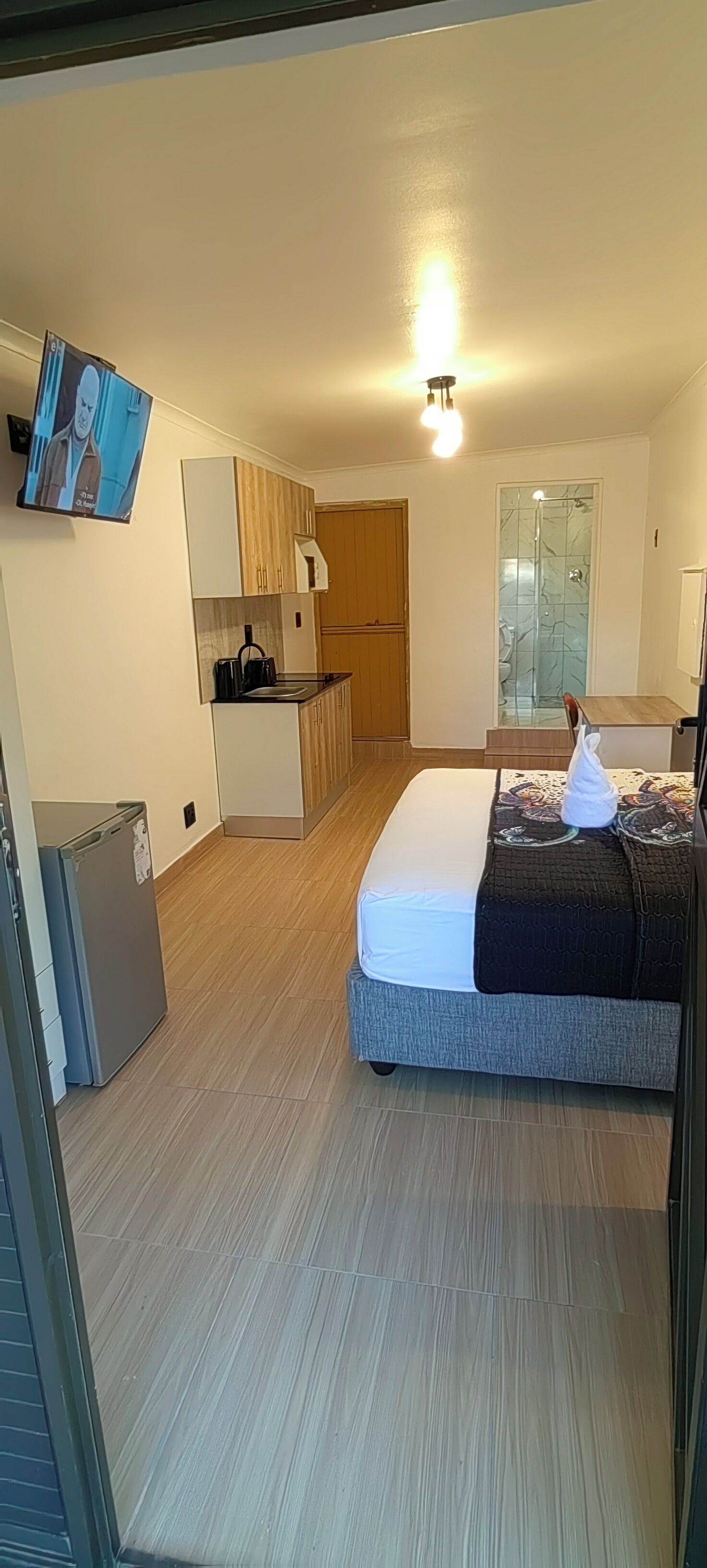 Une chambre compacte comprenant un lit, une petite kitchenette, une douche et un téléviseur fixé au mur.