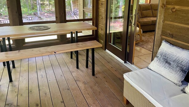 Ett trädäck med en bänk och ett bord, en skjutdörr i glas som leder ut till ett skogsområde och en soffa i rotting med en kudde.