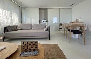 A modern living room with a sofa, a dining table with chairs, a kitchen in the background, and a large window. - Romantic Beachfront Suite with Ocean Views (El Campello)
