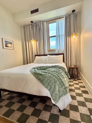 Une chambre avec un sol à carreaux, un lit recouvert de draps blancs, une couverture verte et deux lampes de chevet.