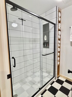 Une douche fermée par des parois de verre, avec un cadre noir, des murs recouverts de carreaux blancs et un sol à carreaux.