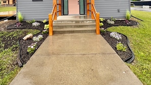 Une maison à deux étages avec un porche à l’avant, un escalier en béton menant à la porte et un petit jardin agrémenté de différentes plantes.