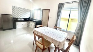 Uma sala de jantar com mesa, cadeiras, geladeira, espaço de cozinha e janela com cortinas.
