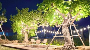 Área externa com assentos, cadeiras e mesas sob uma árvore, decorada com luzes.