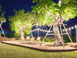 Un área de estar al aire libre con sillas y mesas bajo un árbol, rodeada por una guirnalda decorativa de luces.