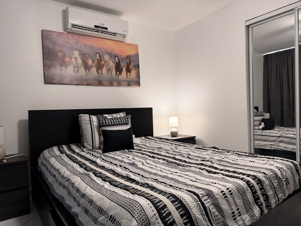 A bedroom with a large bed, a nightstand, a bedside lamp, a wall-mounted air conditioner, and a painting of horses. - My Paradise: Tropical, 3-star, beach, sand—everything at your fingertips (Carolina Puerto Rico)