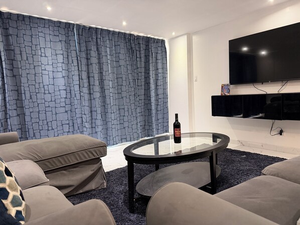A modern living room with a glass coffee table, a flat-screen TV mounted on the wall, and a patterned curtain. - My Paradise: Tropical, 3-star, beach, sand—everything at your fingertips (Carolina Puerto Rico)