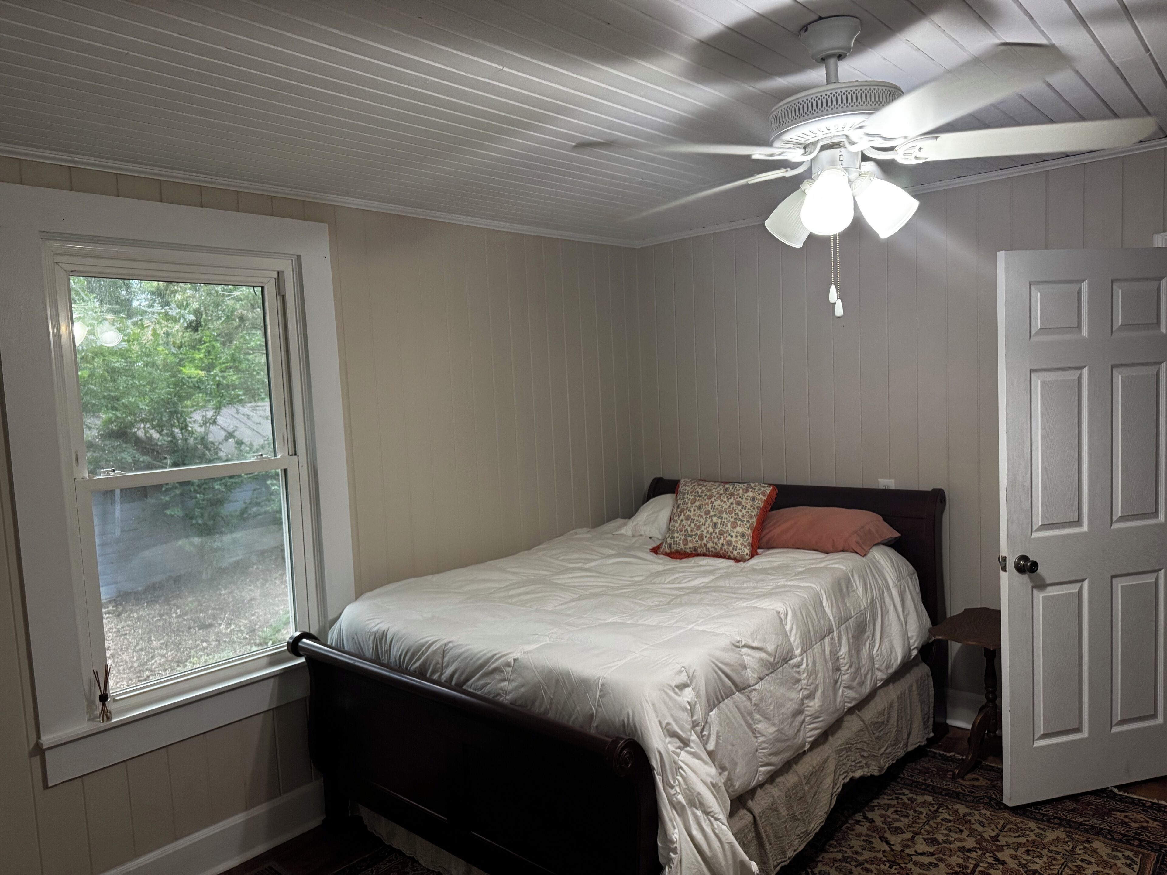 Um quarto com cama, ventilador de teto, janela e porta.