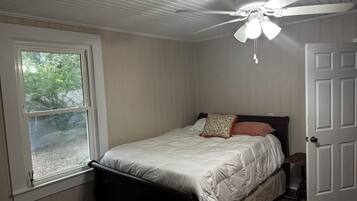 Um quarto com cama, ventilador de teto, janela e porta.