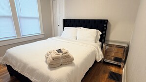 En pænt redt seng med hvide sengetøj, en sort sengegavl og en stak foldede håndklæder for foden af sengen.