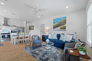 A modern living room with a blue sofa, dining area with white chairs, and a kitchen with stainless steel appliances. - Bayou Breeze  - Pool + Waterfront Views + 8 Minutes to Downtown Pensacola (Pensacola)