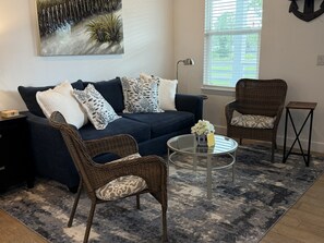 A living room with a dark blue sofa, patterned cushions, a wicker chair, a glass coffee table with a vase, and a window with blinds. - Bayou Breeze  - Pool + Waterfront Views + 8 Minutes to Downtown Pensacola (Pensacola)