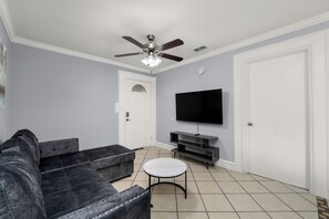 Ein Wohnzimmer mit einem dunklen Sofa, einem runden Couchtisch, einem Deckenventilator, einem Flachbildfernseher und einer weißen Tür.