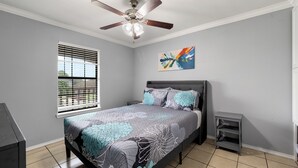 A bedroom with a bed, ceiling fan, window with blinds, artwork on the wall, and a small bedside table. - The C4 Gateway to Starbase (Brownsville)