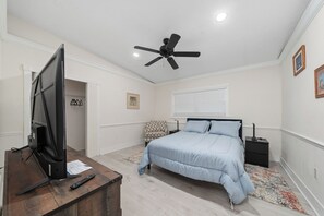 Een slaapkamer met een bed, een plafondventilator, een tv op een houten standaard, een nachtkastje en een raam met jaloezieën.