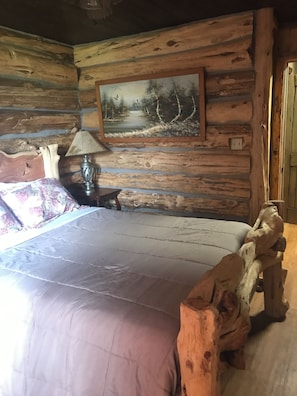 Una habitación tipo cabaña de madera con una cama, una lámpara, un cuadro de un paisaje forestal y un cabecero de madera.
