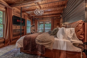 Ein Schlafzimmer in einer Holzhütte mit einem Bett, einem Fernseher, einem Hängesessel und Blick ins Freie.