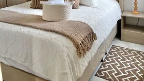 A neatly made bed with a beige headboard, a bedside table with a lamp, and a patterned rug on the floor.
