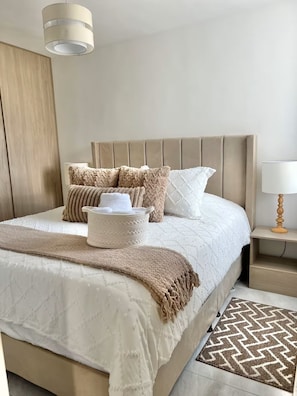 A neatly made bed with a beige headboard, a bedside table with a lamp, and a patterned rug on the floor.