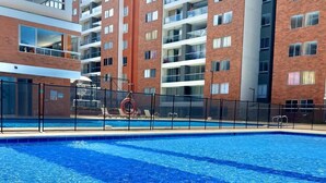 A swimming pool with a clear blue water surface, surrounded by a black fence. In the background, there are multi-story buildings with balconies and windows.