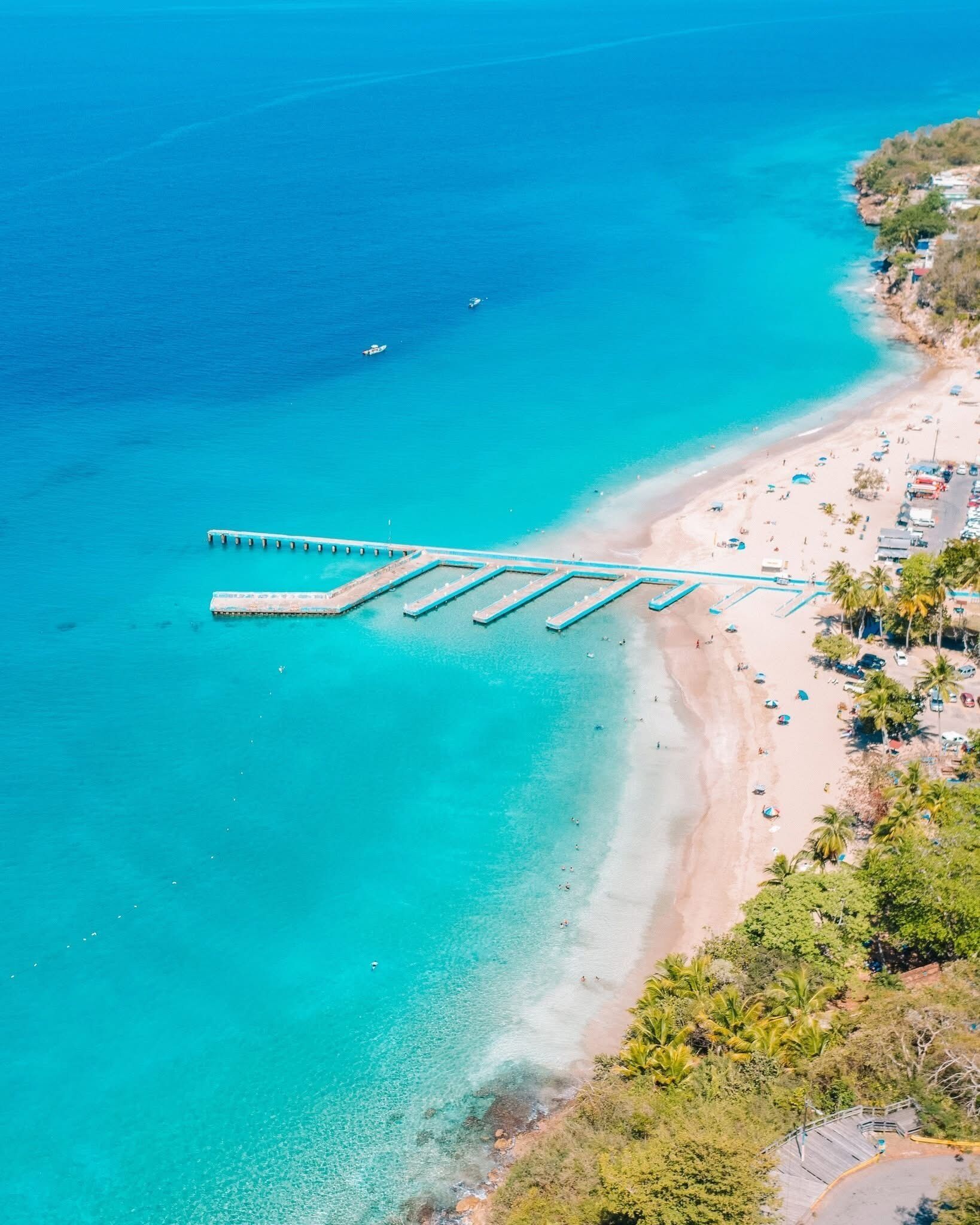 An aerial view of a beach with a pier extending into the water, surrounded by clear turquoise waters and a sandy shore.