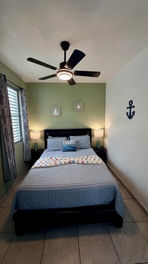 Ein Schlafzimmer mit einem Bett, einem Deckenventilator, zwei Nachttischlampen, einem Fenster mit Vorhängen und einer Wand mit einer nautischen Anker-Dekoration.