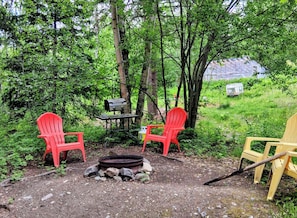 Un espace extérieur aménagé avec deux chaises rouges, une table et un barbecue, le tout entouré d’arbres et agrémenté d’un foyer.
