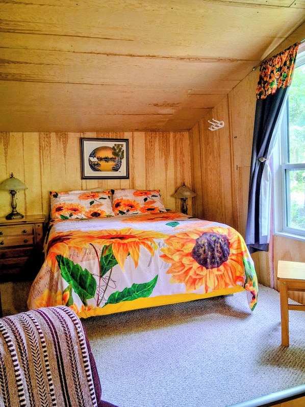 Une chambre à coucher avec un lit, un tableau encadré au mur, une fenêtre avec des rideaux et une petite chaise en bois.