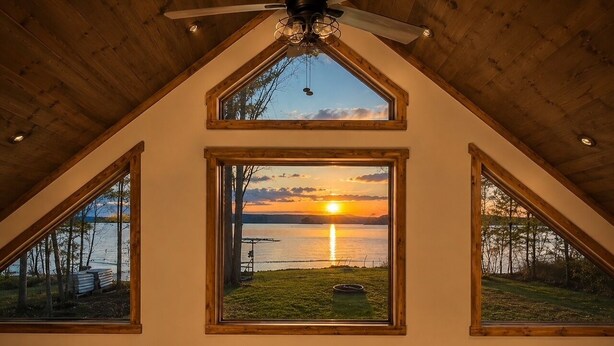 Une pièce avec un plafond en pente, de grandes fenêtres et une vue sur un coucher de soleil sur l’eau.
