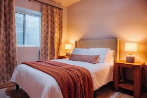 A neatly made bed with a brown blanket, two bedside lamps, and a window with curtains. - Cozy Lakefront Retreat | Sauna, Hot Tub & Ski (Wild Rose)
