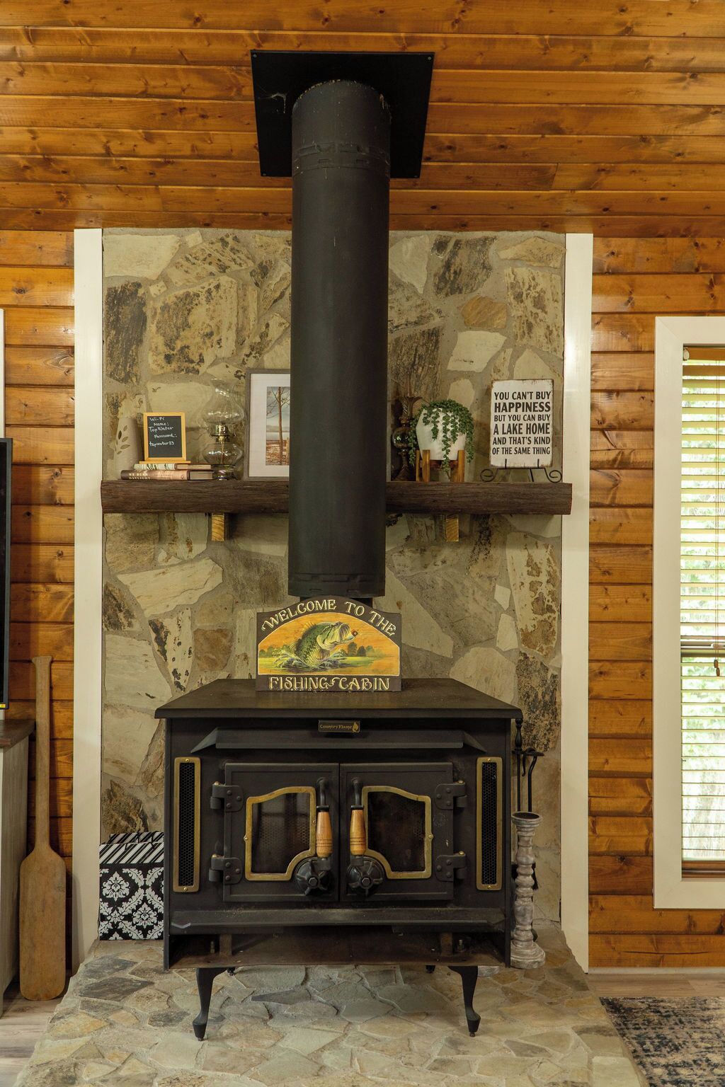 Ein Kaminofen mit einem Schild 'Willkommen in der Fischerhütte', Steinwänden, Holzregalen mit Büchern und einer Pflanze sowie einem Steinfußboden.